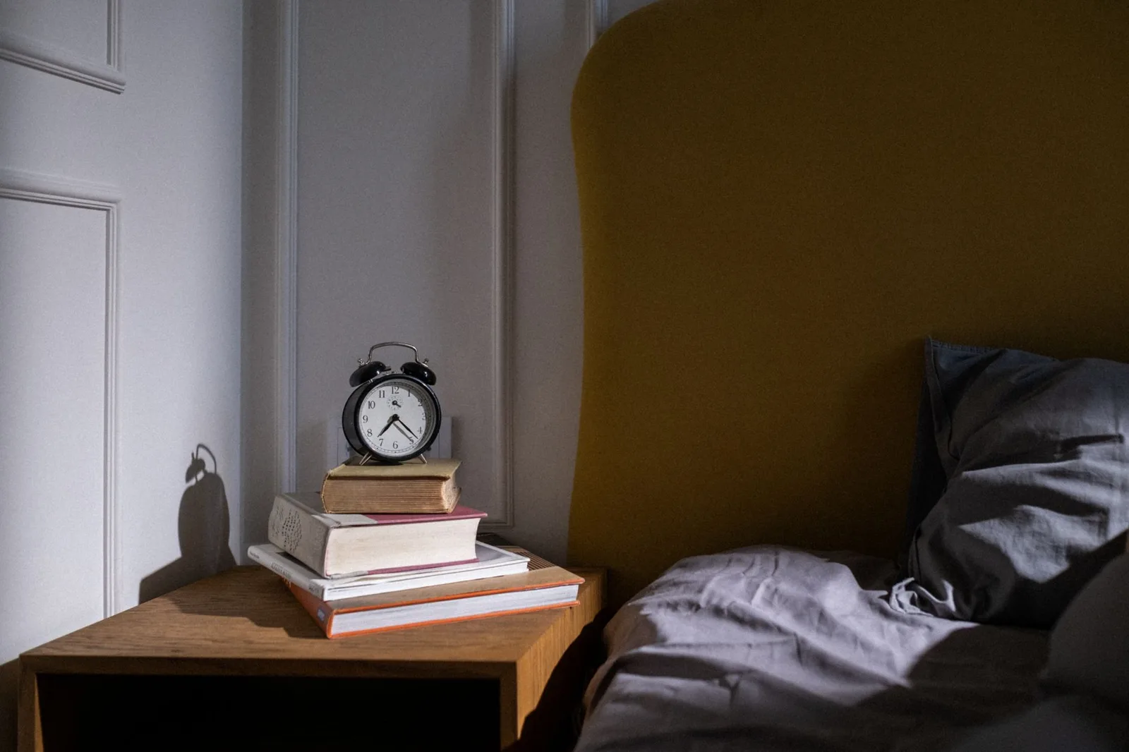 Bedside table with an alarm clock and a stack of books next to a rumpled bed